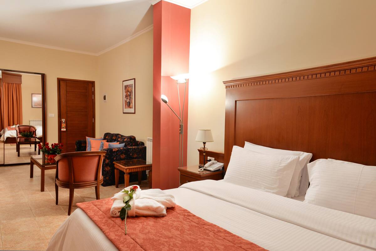 A hotel room with a large bed, a sitting area with a sofa and armchairs, a wooden headboard, a nightstand with a lamp, and a bathroom visible through a door.