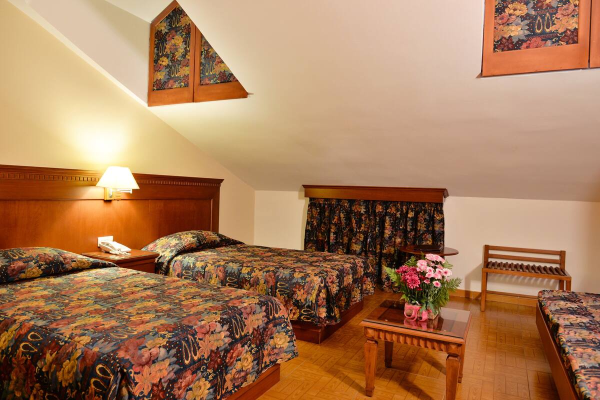 A hotel room with two beds, a wooden headboard, a nightstand with a lamp, a small table with a vase of flowers, and a wooden bench.