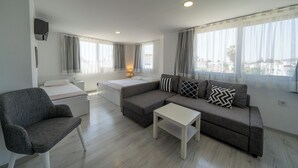 Una sala de estar moderna con un sofá gris, una mesita de centro blanca y un sillón gris. Se ve una cama con ropa de cama blanca al fondo.