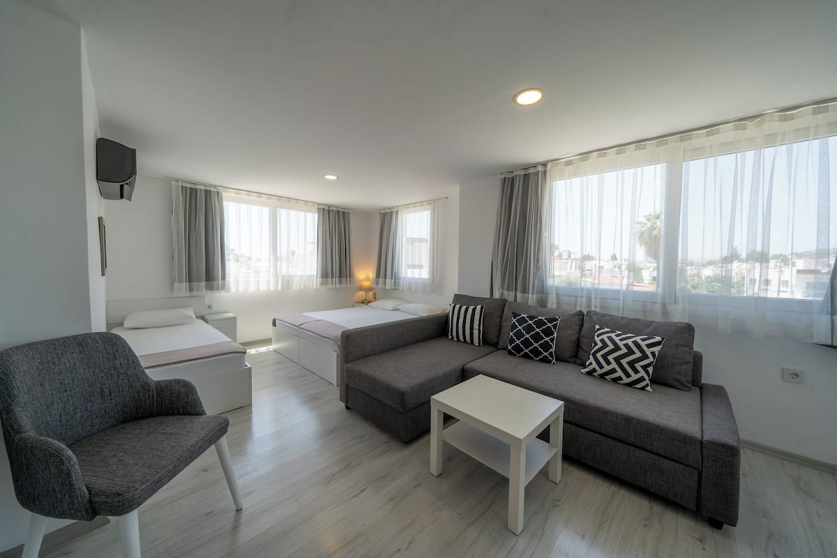 Una sala de estar moderna con un sofá gris, una mesita de centro blanca y un sillón gris. Se ve una cama con ropa de cama blanca al fondo.