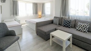 Una sala de estar moderna con un sofá gris, una mesa de centro blanca y un sillón gris. Hay dos camas con ropa de cama blanca y una mesita de noche con una lámpara.