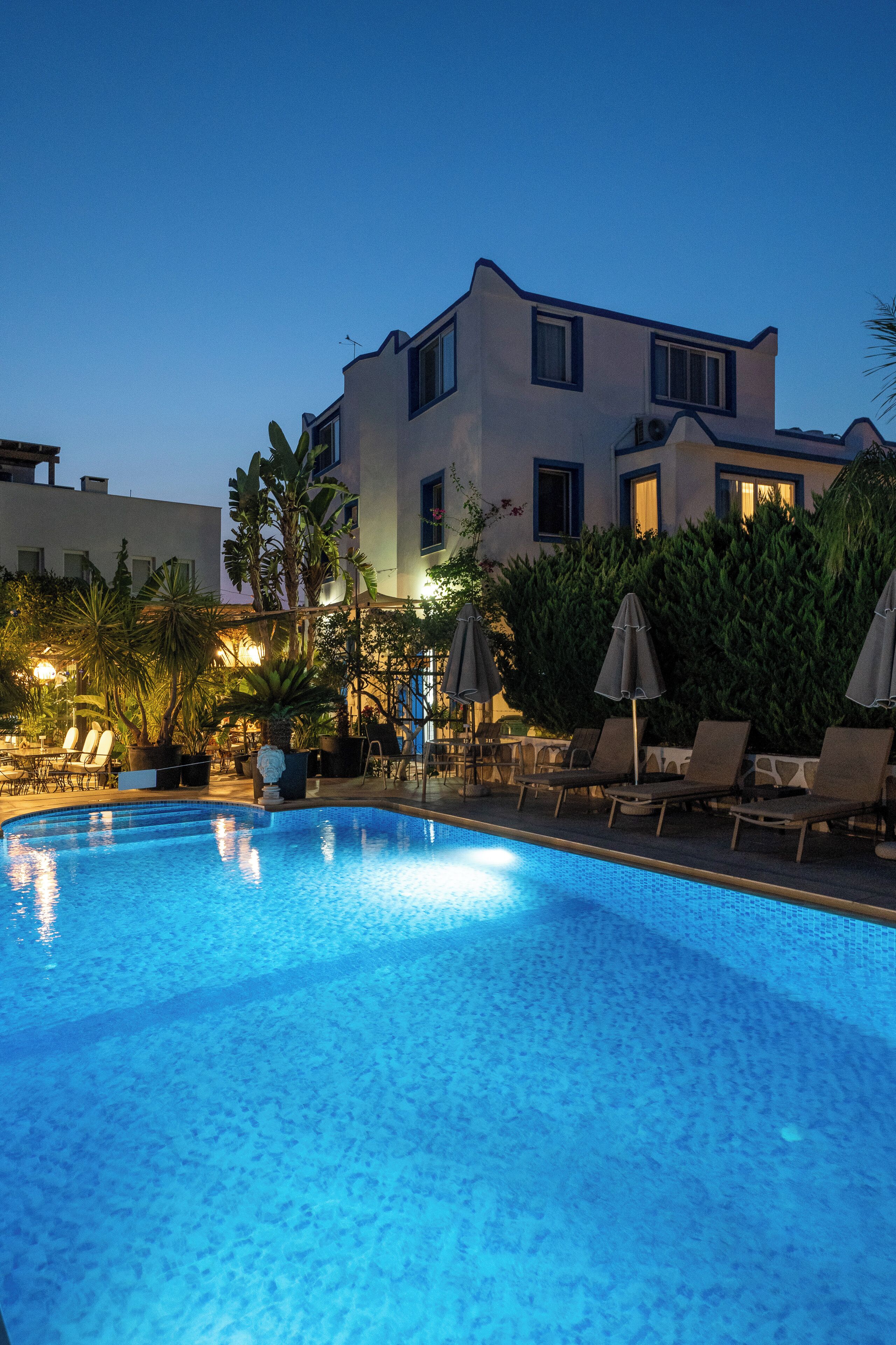 A swimming pool with lounge chairs and umbrellas, surrounded by greenery and a building with multiple windows.