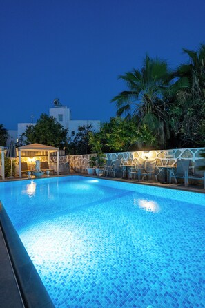 Una piscina con piso de baldosas, rodeada de sillas de descanso y una zona de estar cubierta con iluminación ambiental.