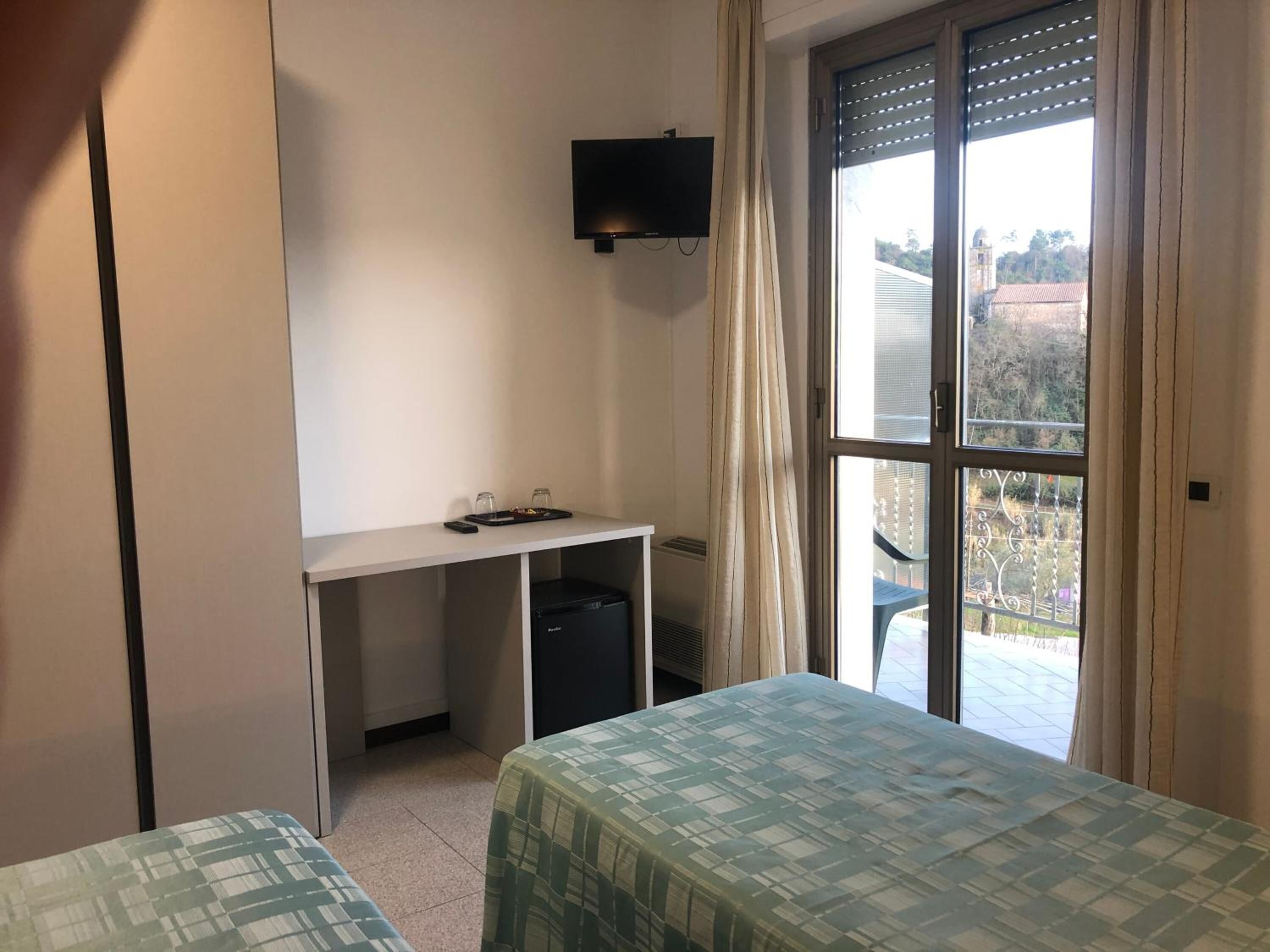 Una stanza piccola con due letti, una scrivania, una TV e una finestra che si affaccia su alberi ed edifici.