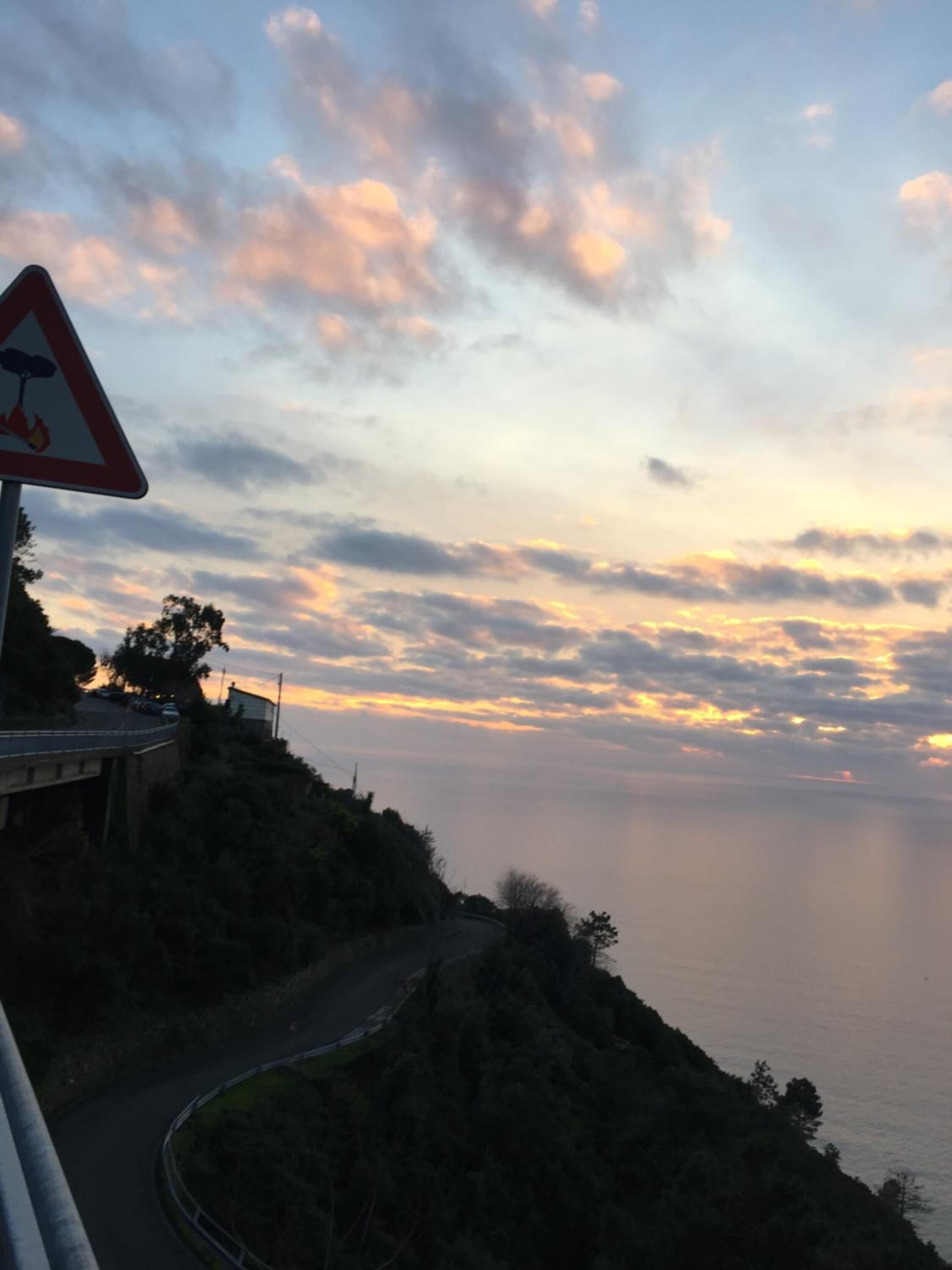 Une route côtière, agrémentée d’un panneau d’avertissement concernant les risques d’incendie, bordée d’une végétation luxuriante et offrant une vue panoramique sur un coucher de soleil magnifique.