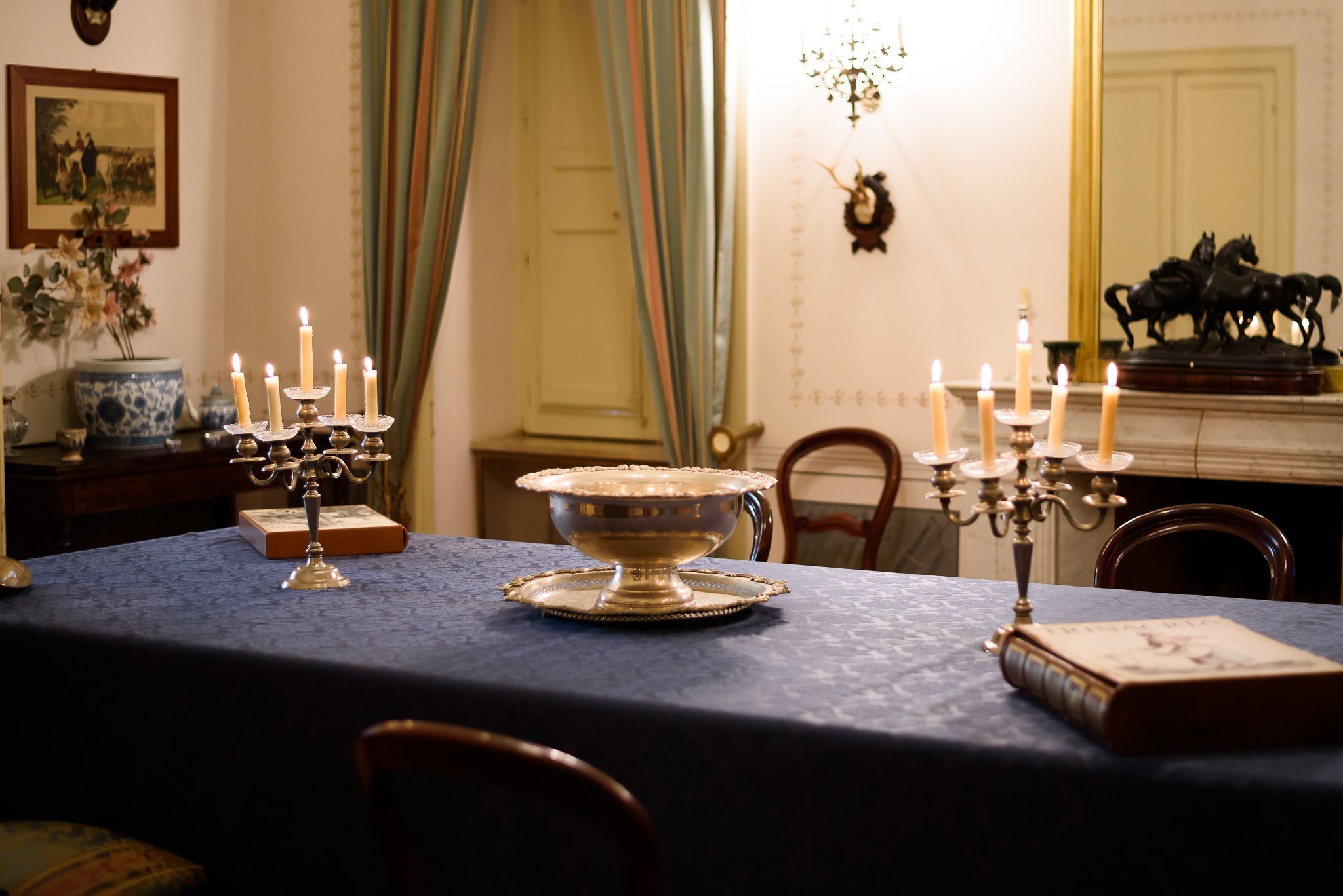 A dining table with a blue tablecloth, a silver candelabra with lit candles, a decorative bowl, and a book.