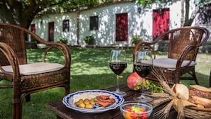 Un espacio exterior para comer con sillas de mimbre, una mesa con comida y bebidas, y un edificio blanco al fondo.