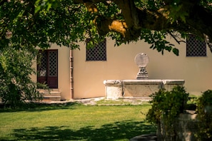 Un banco de piedra con un adorno superior, un edificio con puerta y ventanas, y un árbol en primer plano.