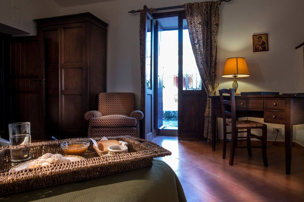 Una habitación con una cama, una bandeja de mimbre con alimentos para el desayuno, una silla, un escritorio y una lámpara.