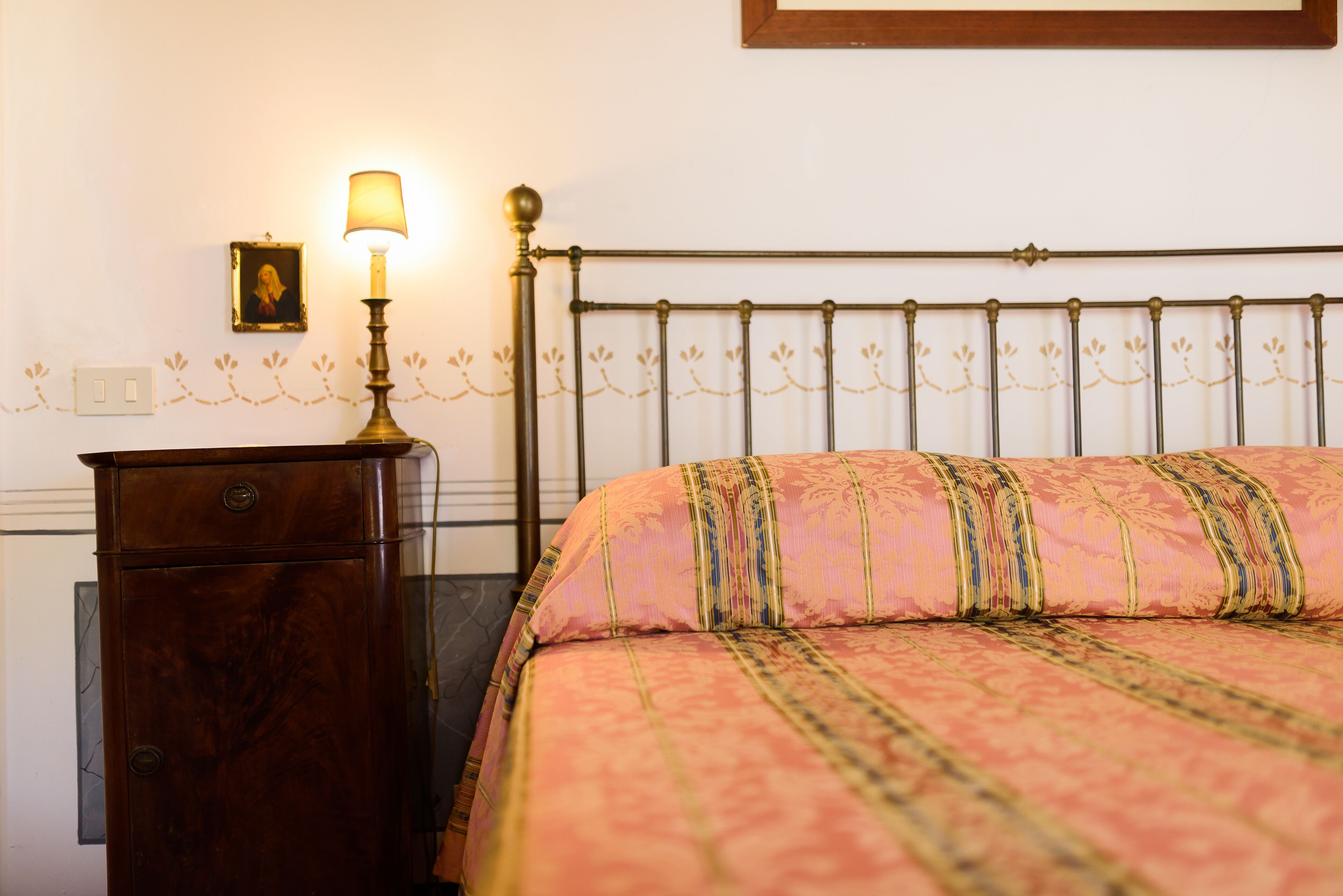 A bed with a metal headboard, a wooden nightstand with a lamp, and a framed picture on the wall.