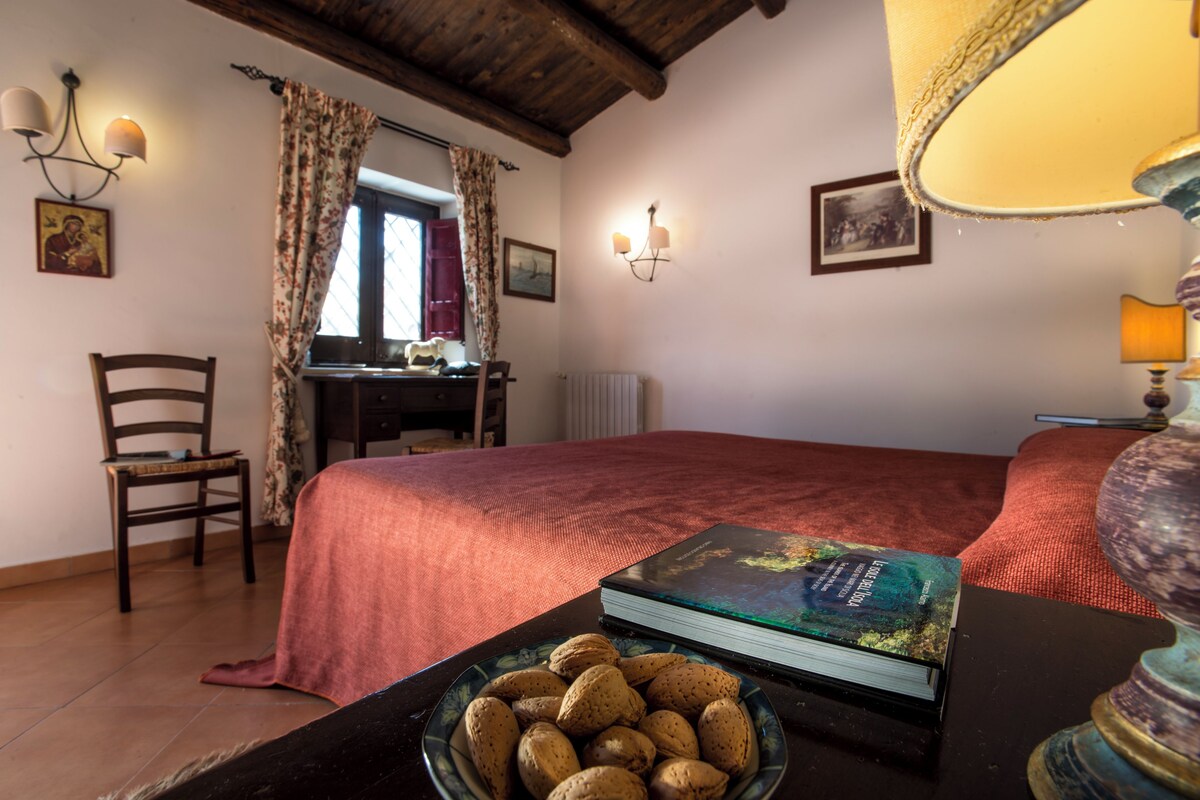 Un dormitorio con cama, una silla, una mesa con un libro y un cuenco de nueces, una lámpara y una ventana con cortinas.