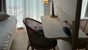 A hotel room with a bed, a desk with a lamp, a chair, a TV mounted on the wall, and a view of the sea through sheer curtains. - Les Sables Blancs (Concarneau)