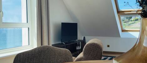 Een kamer met een ronde houten tafel, twee gestreept stoelen, een televisie op een standaard en een raam met gordijnen.