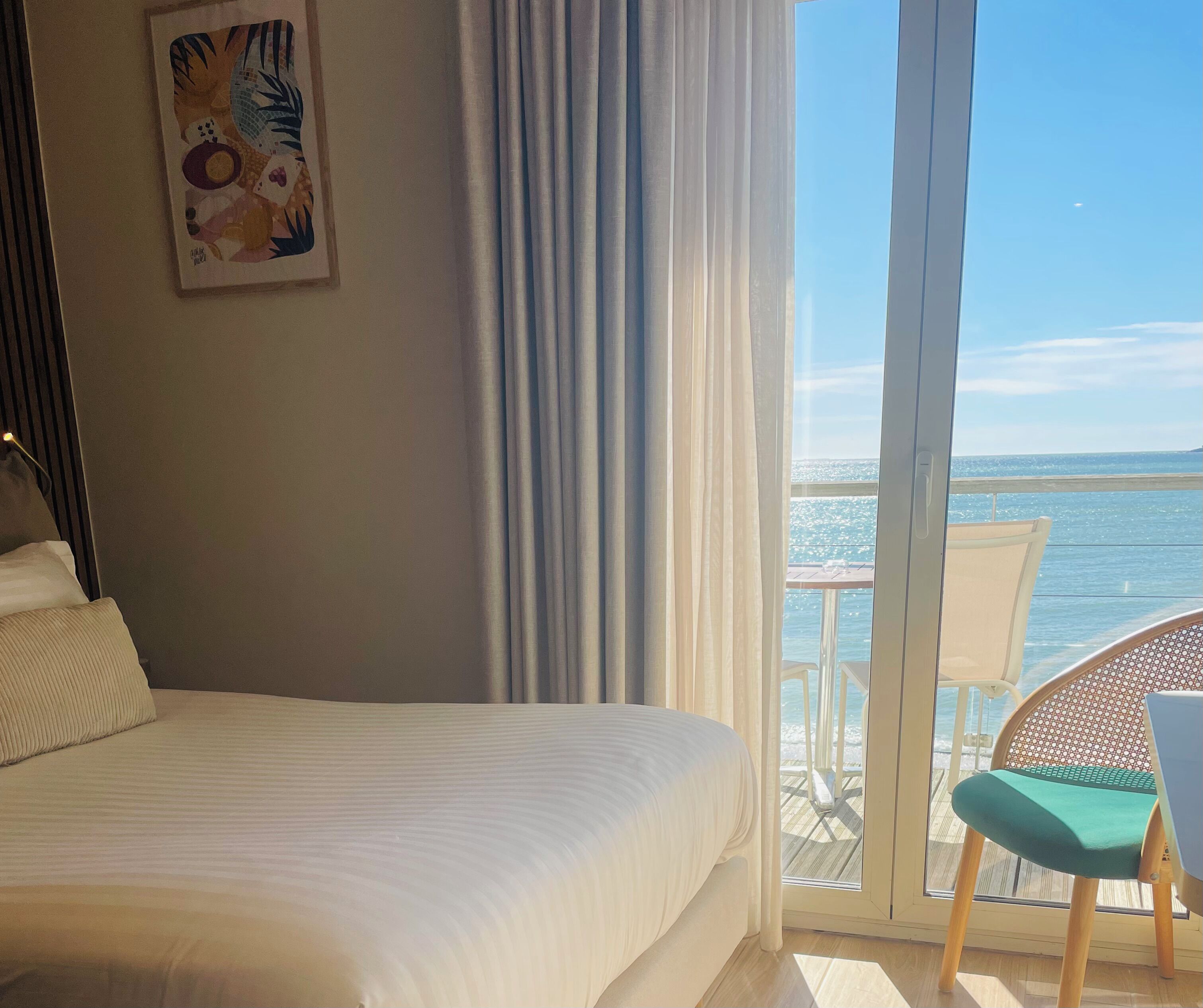 A hotel room with a bed, a painting on the wall, and a view of the sea through a glass door.