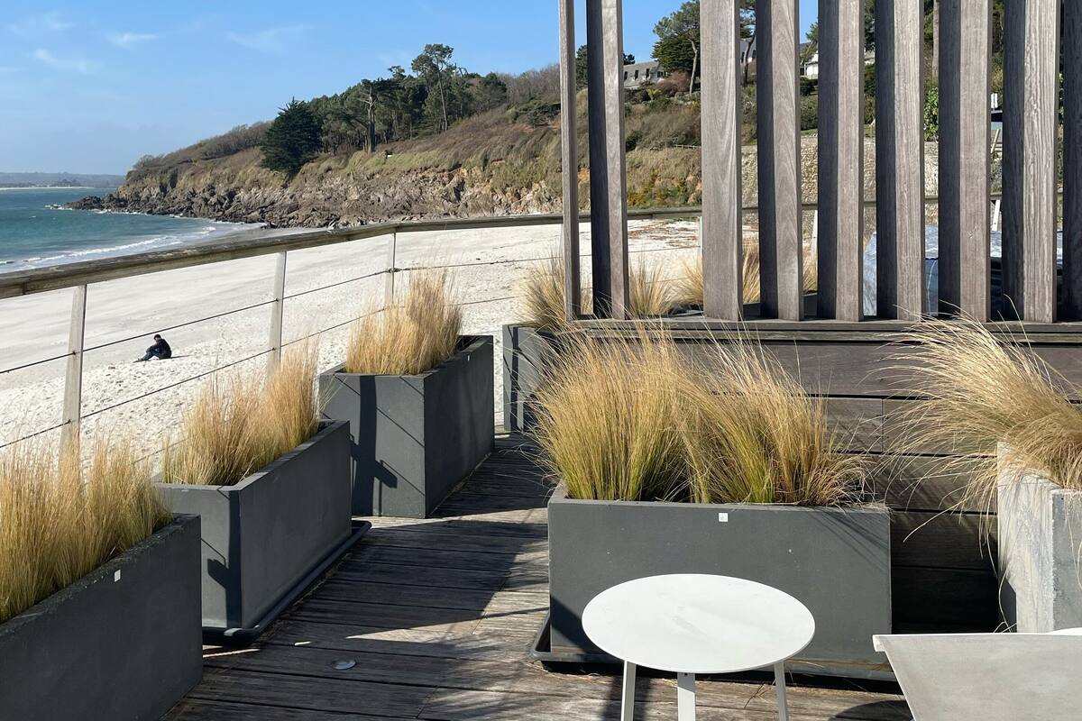 Una terraza de madera con una mesa y sillas blancas, con vista a una playa donde hay una persona sentada en la arena.