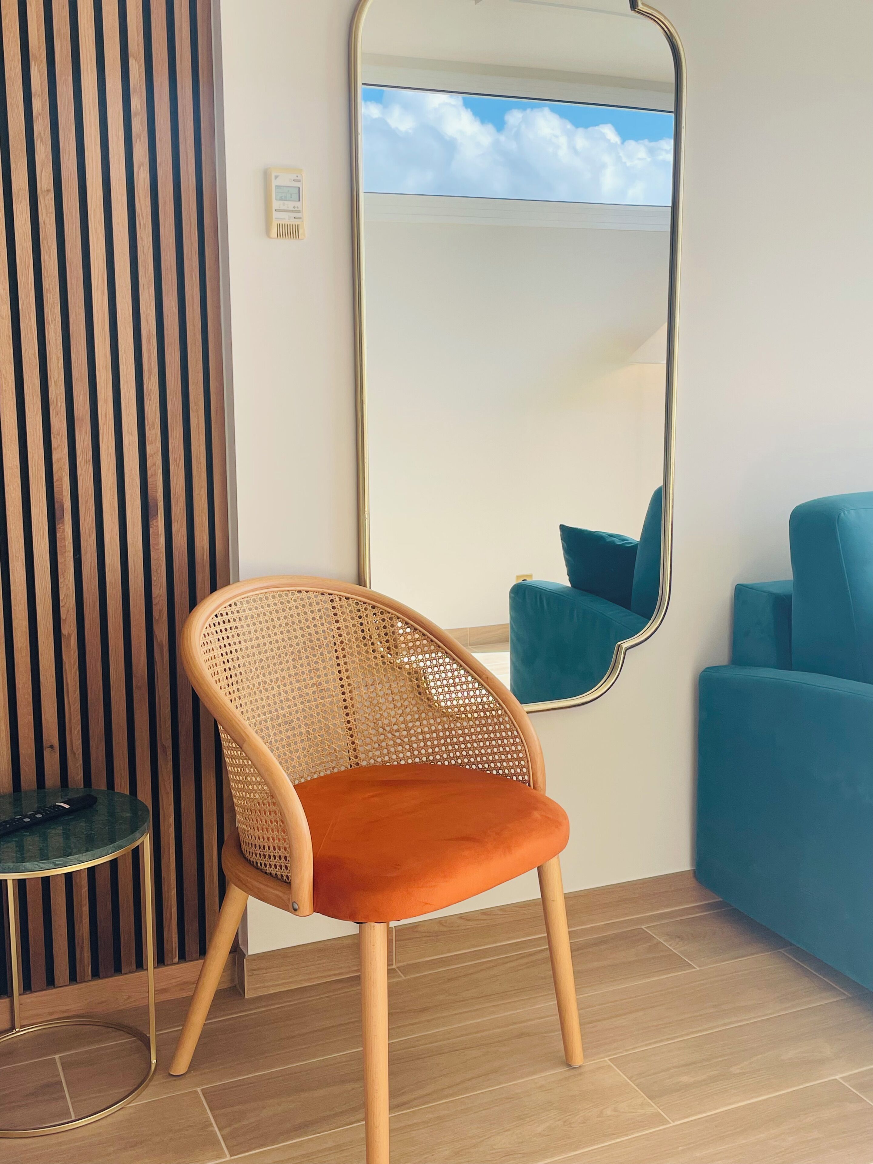 A room with a large mirror, a wicker chair with orange cushions, a small round table, and a blue sofa.