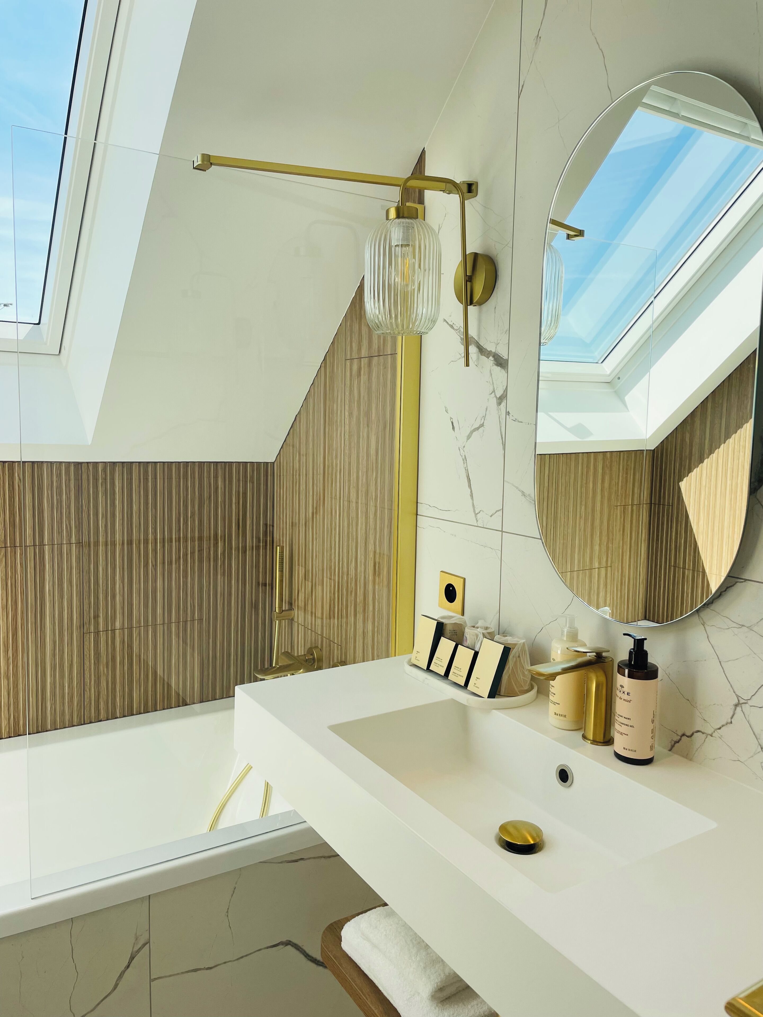 A modern bathroom with a skylight, a large mirror, a white sink, and a gold faucet.