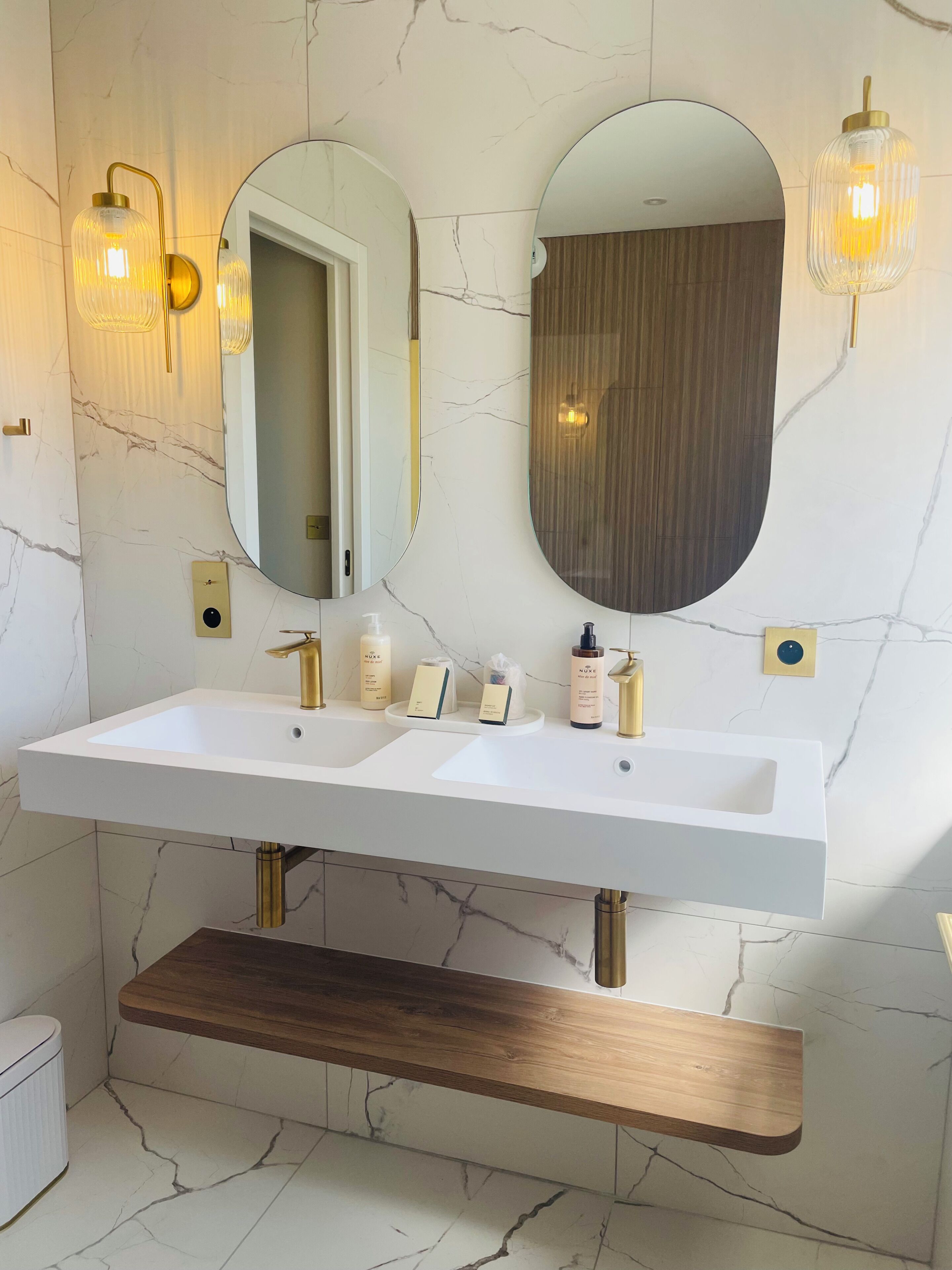 Een badkamer met twee wasbakken, een spiegel, wandlampen en een houten plank.