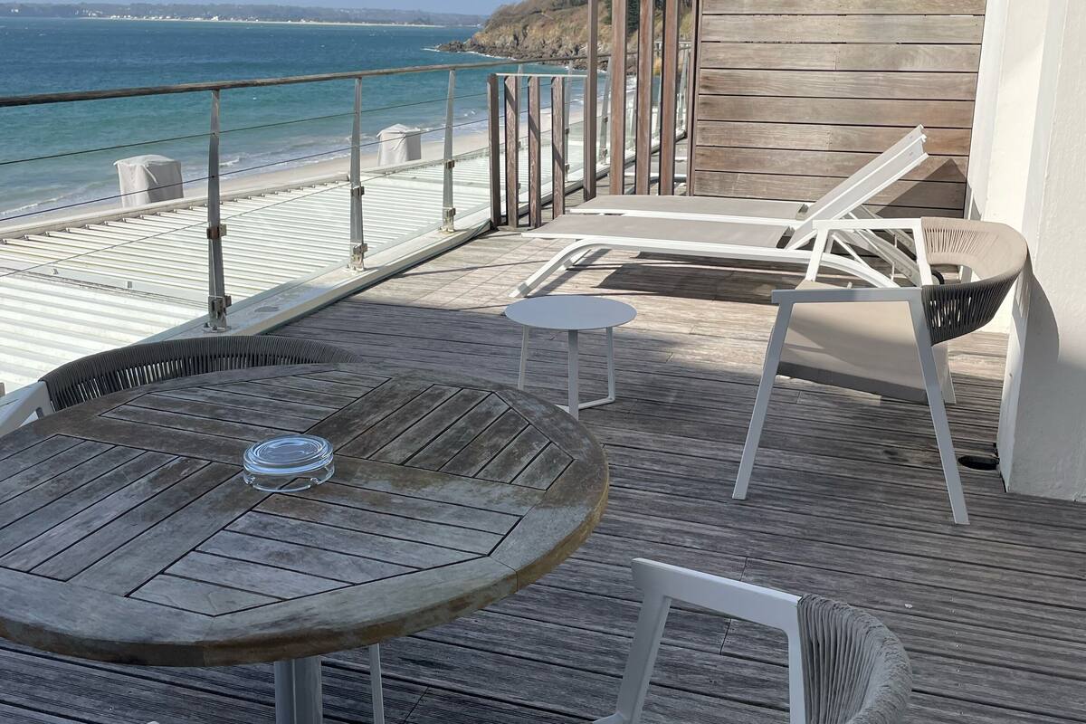 Una terraza de madera con una mesa redonda y sillas, con vista a una playa y el océano.