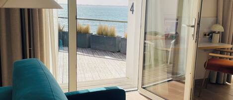 Una sala de estar moderna con un sofá color turquesa, un ventanal de vidrio que da a un balcón con vistas al mar, y un televisor de pantalla plana montado en la pared.