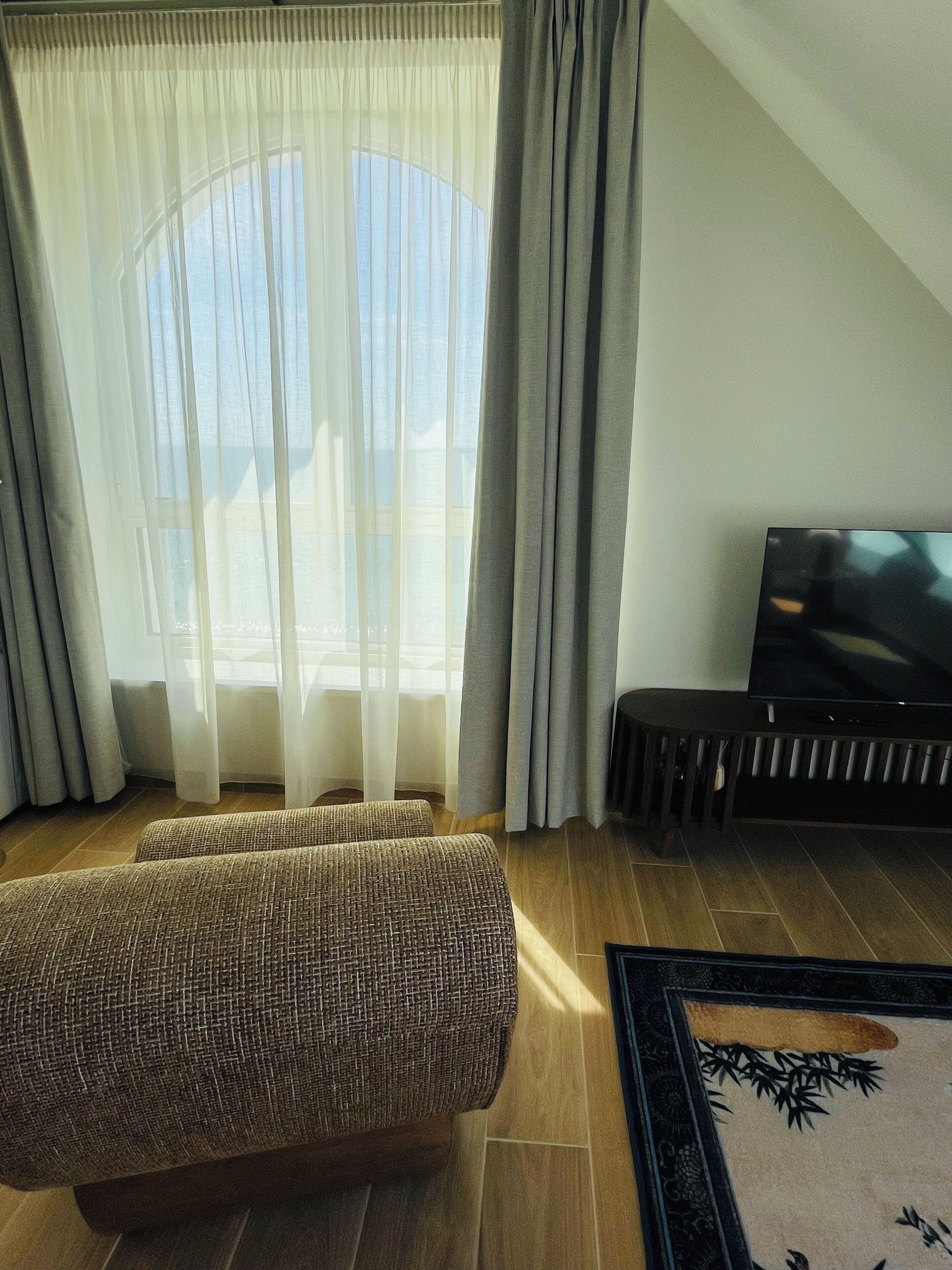 Una sala de estar con un sofá, un televisor sobre un mueble bajo y un ventanal con cortinas traslúcidas.