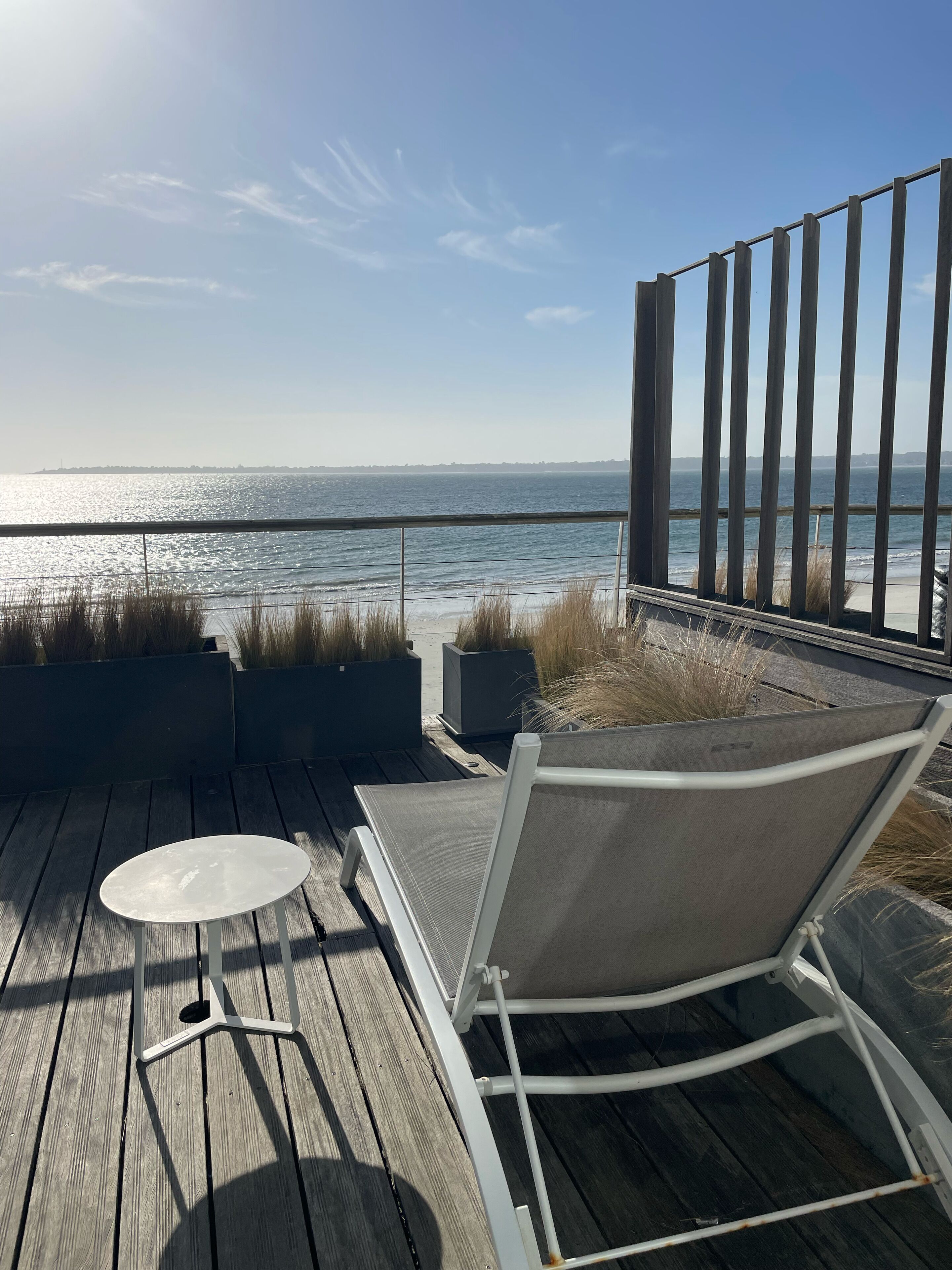 A wooden deck with a white chair and table, overlooking a body of water with a distant shoreline.