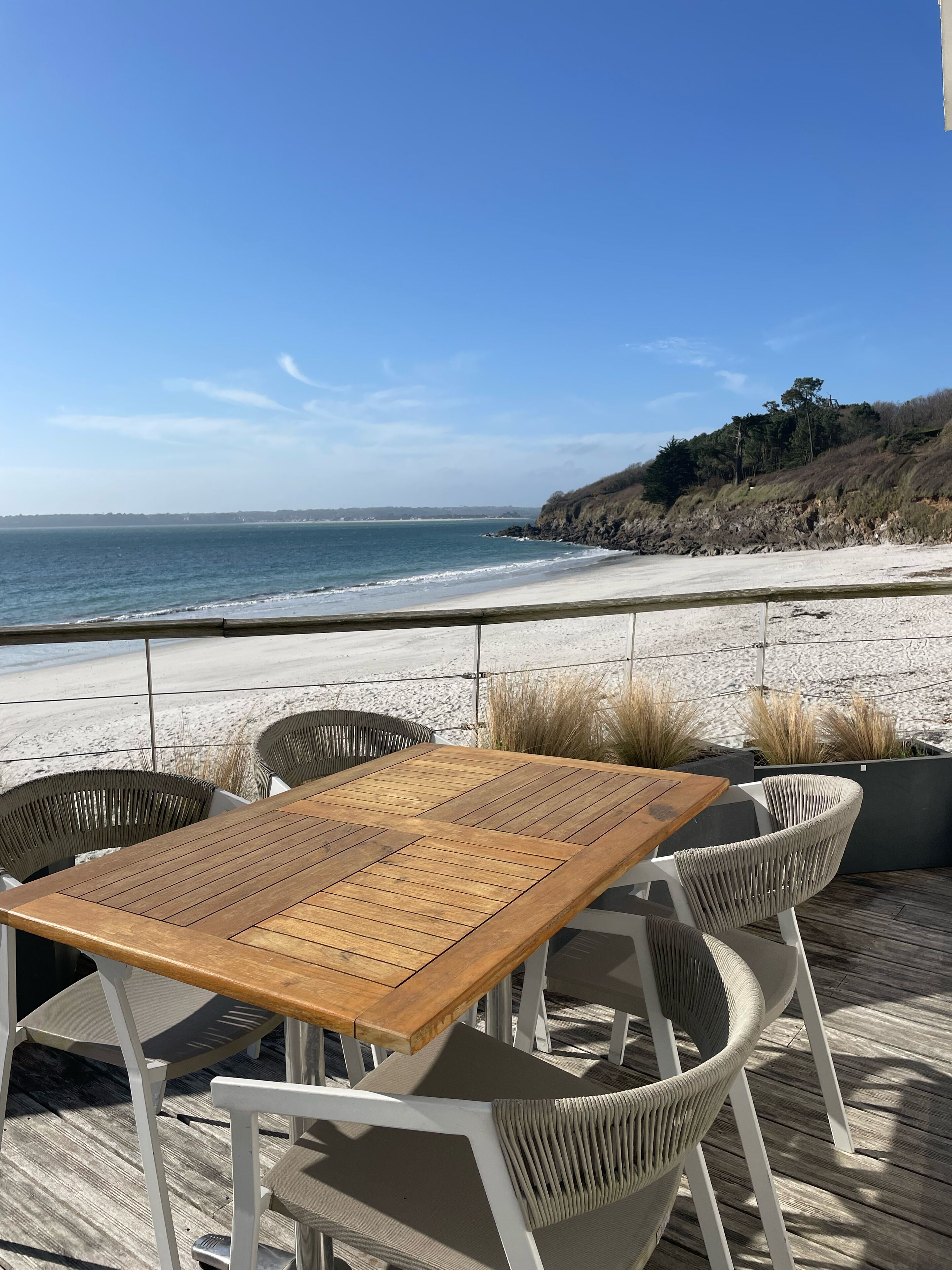 Una mesa de madera y sillas blancas en una terraza con vistas a una playa y al océano.