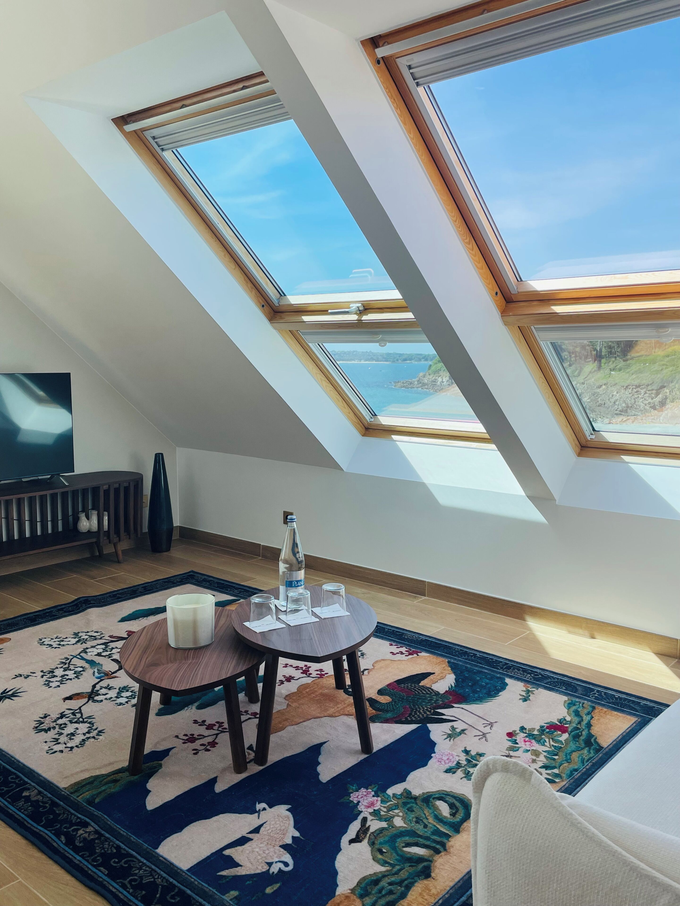 Een kamer met een schuine kap, dakramen, een ronde tafel met een fles en glazen, een televisie en een decoratief vloerkleed.