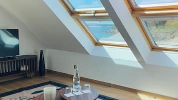Een kamer met een schuine kap, dakramen, een ronde tafel met een fles en glazen, een televisie en een decoratief vloerkleed.