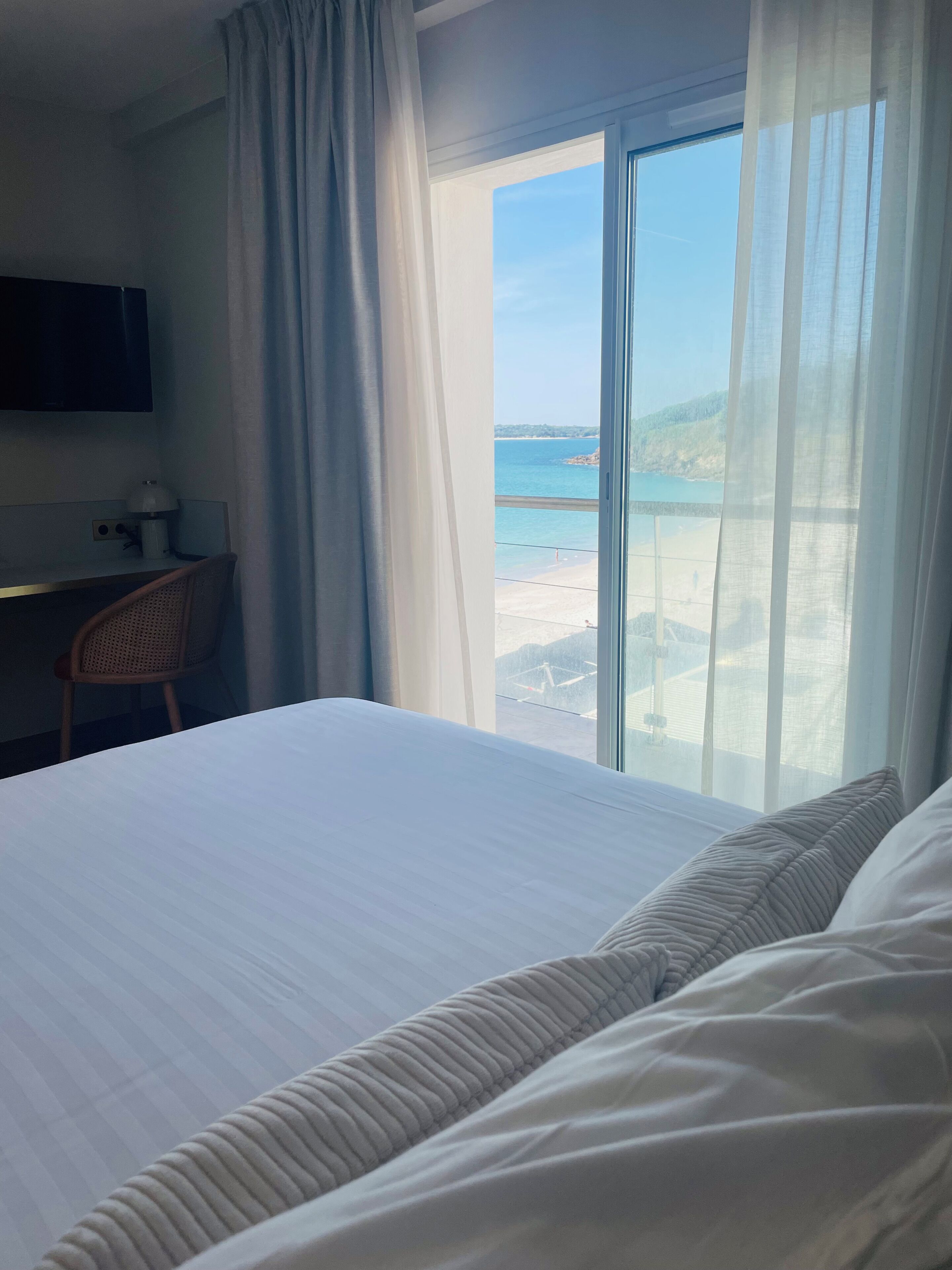 A hotel room with a bed, a desk, a chair, a TV, and a large window with a view of the sea.