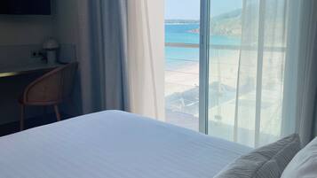 A hotel room with a bed, a desk, a chair, a TV, and a large window with a view of the sea.