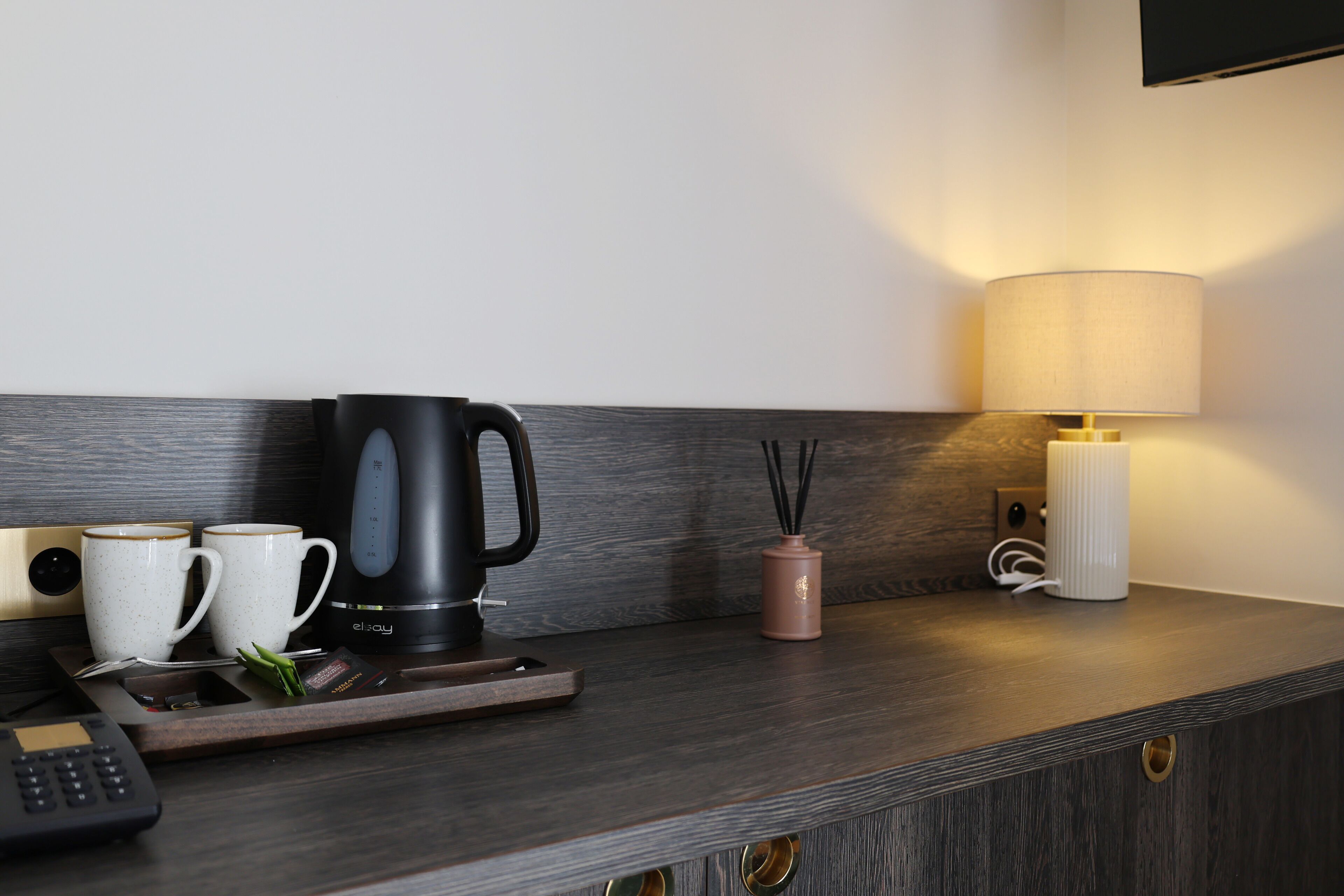 Una habitación de hotel moderna con una encimera de madera oscura, una cafetera negra, dos tazas blancas, un portavelas rosa, una lámpara blanca y una planta pequeña.