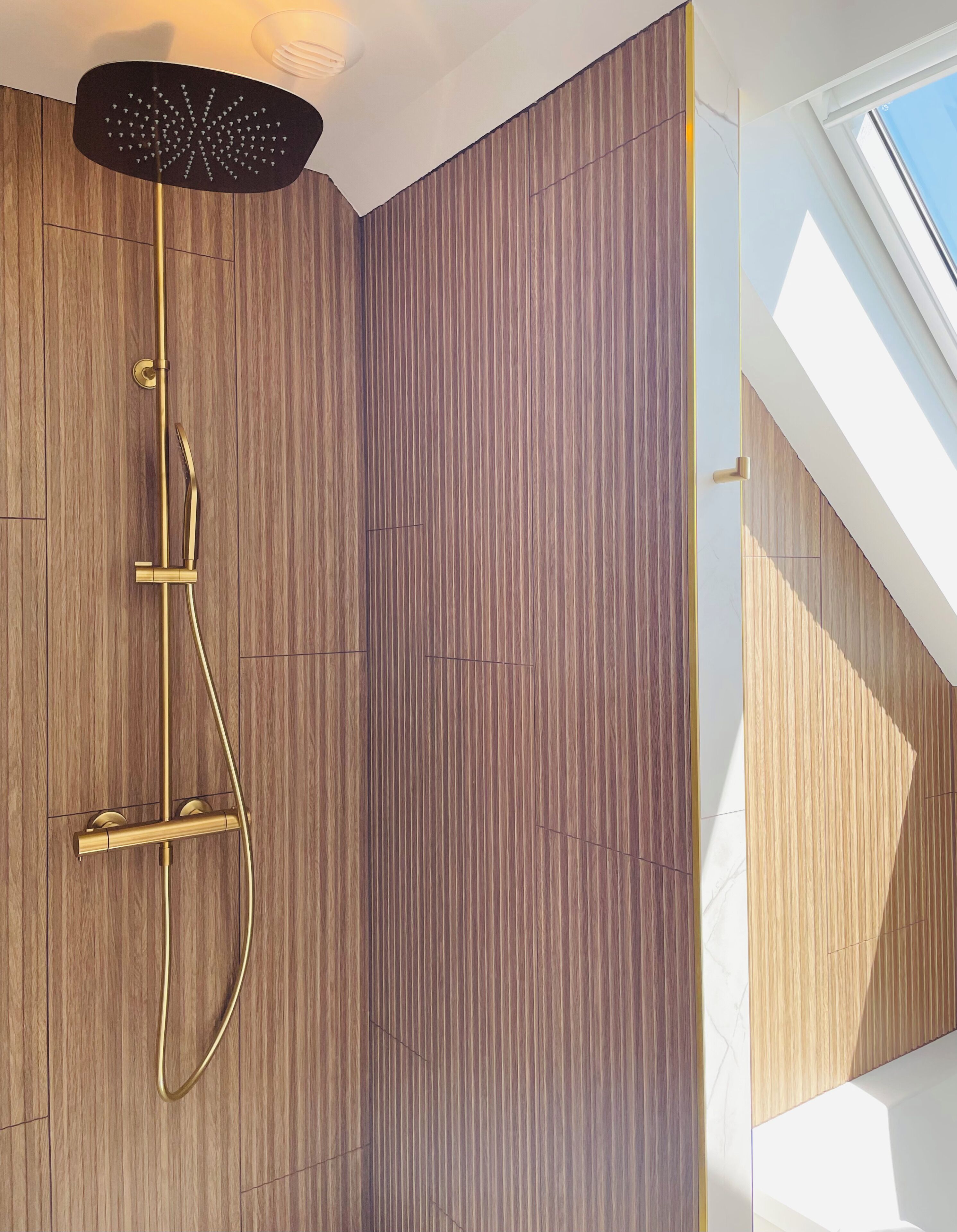 Une douche moderne avec une pomme de douche noire, des accessoires dorés et un mur en bois.