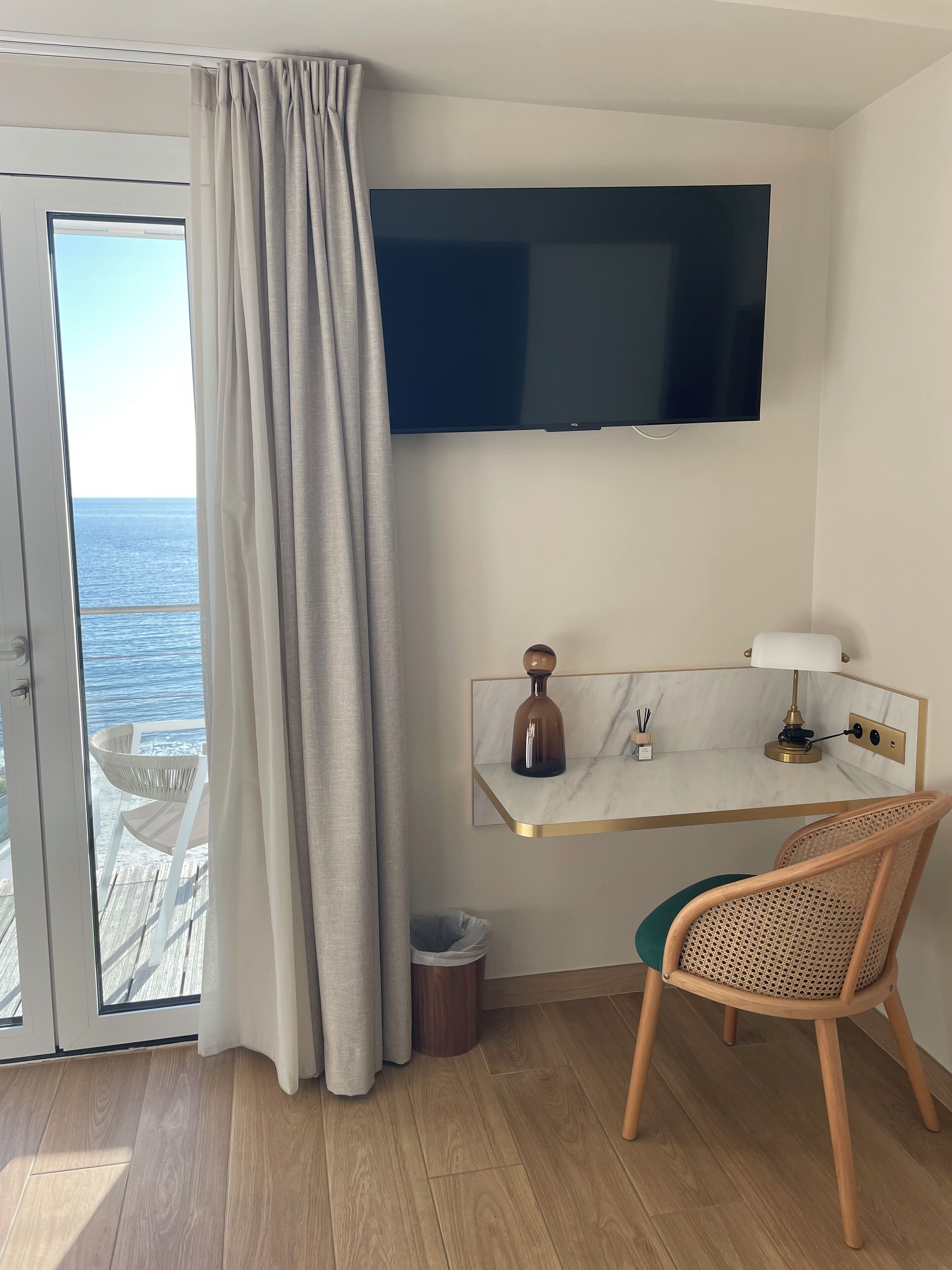 Quarto com televisão na parede, escrivaninha com luminária, uma cadeira, vista para o mar pela janela e uma pequena lixeira.