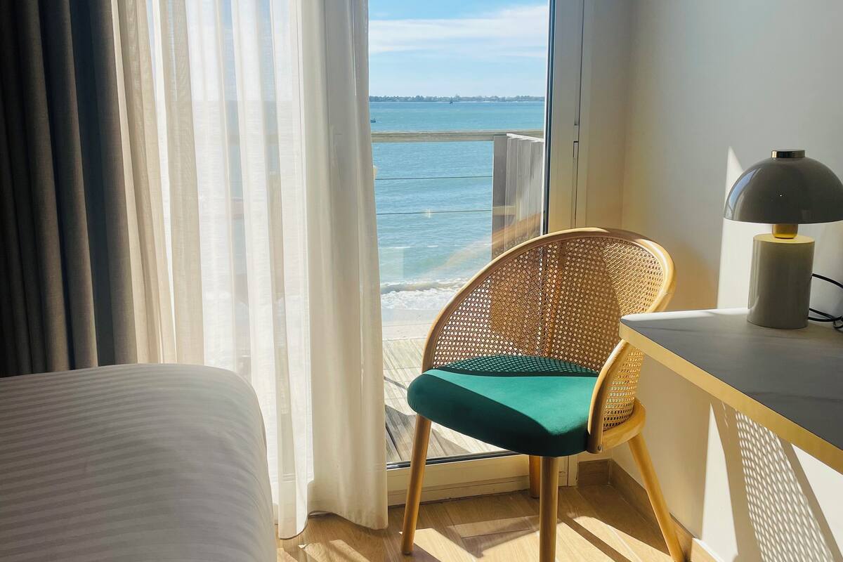 Habitación de hotel con cama, sillón con cojín, escritorio con lámpara y vista al mar a través de una ventana.