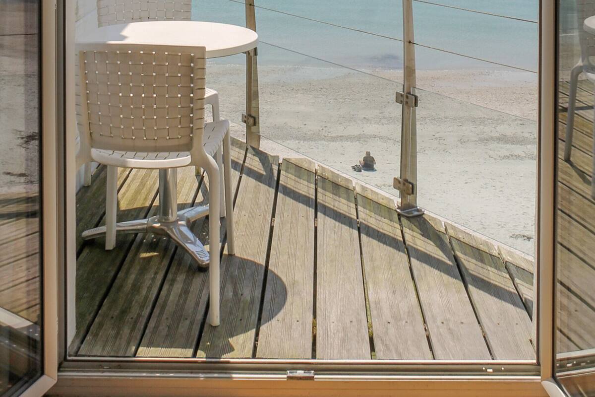 Un balcón con una mesa y una silla blancos, con vista a una playa que tiene un muelle de madera y una barandilla.