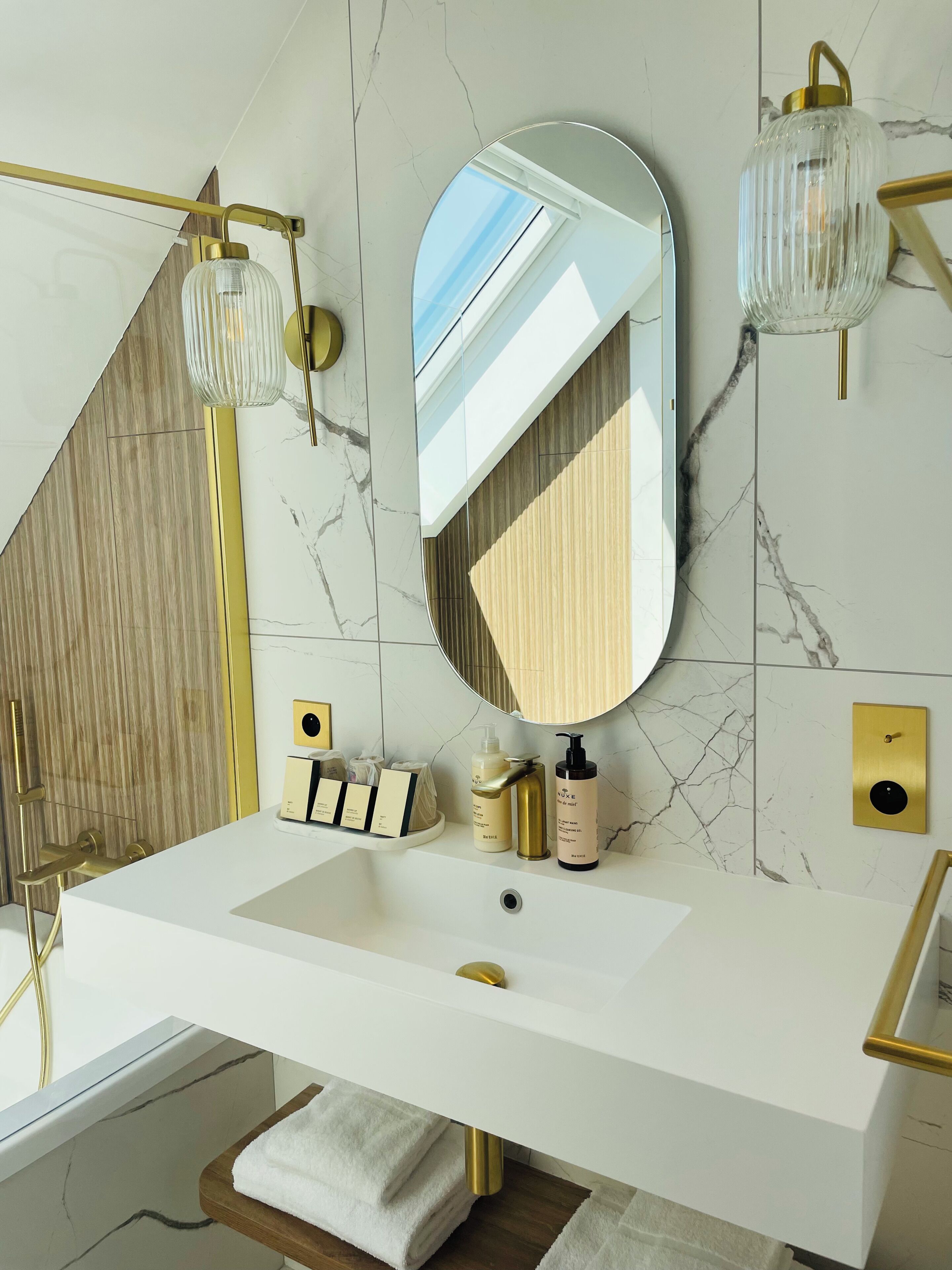 A modern bathroom with a white sink, gold fixtures, a large mirror, and marble walls.