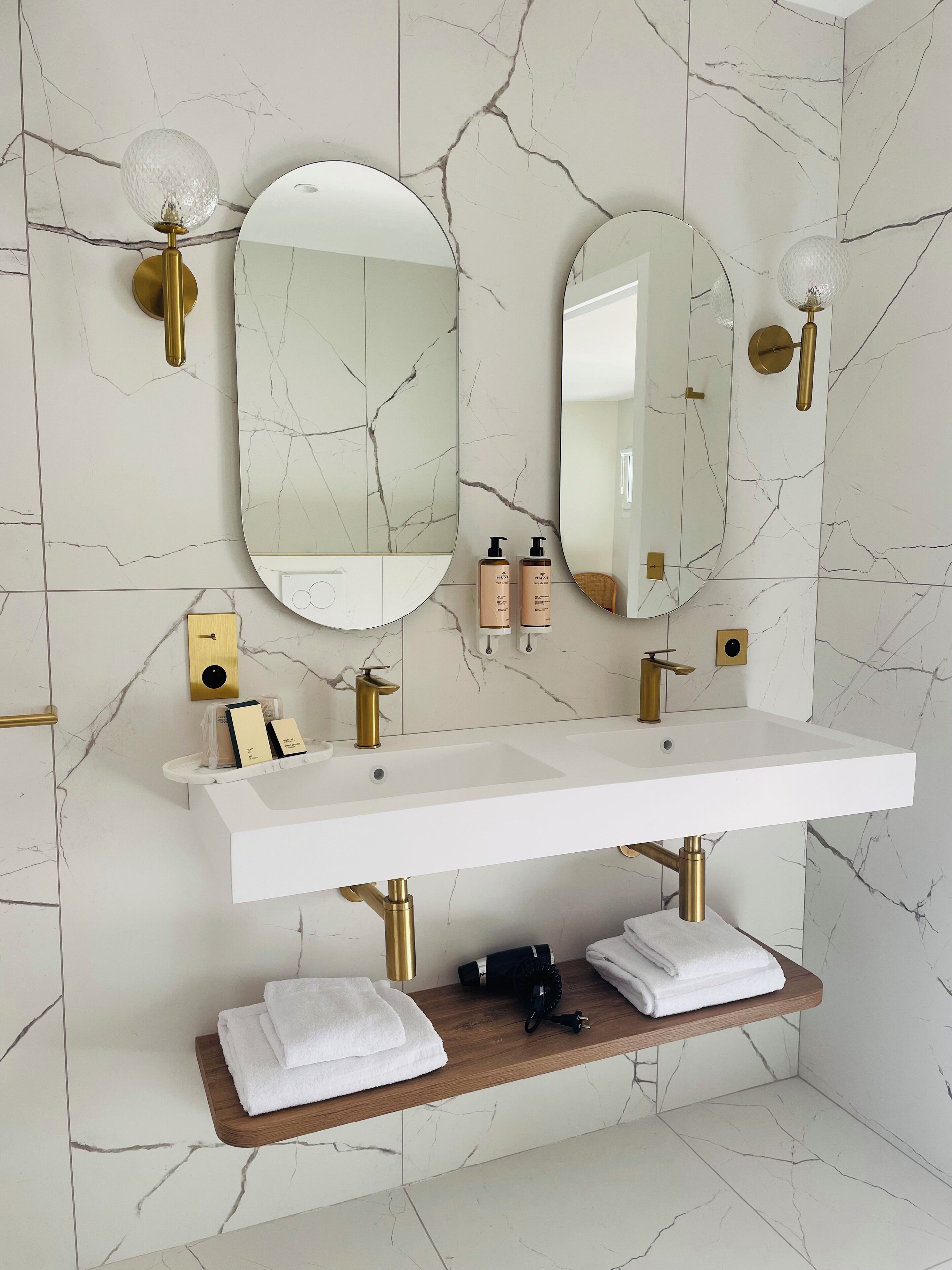 A bathroom with a white marble backsplash, a white sink with gold fixtures, two round mirrors, and a wooden shelf with towels.