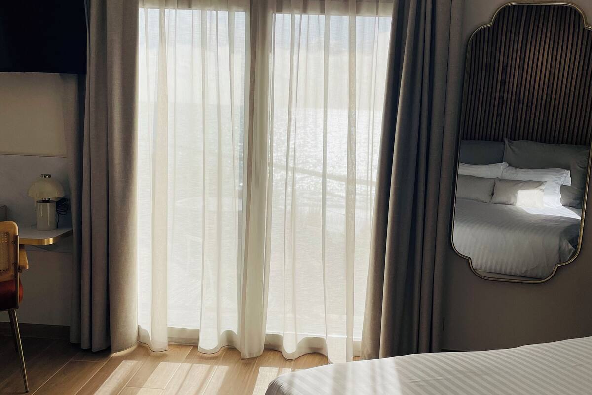 Habitación de hotel con una cama grande, un escritorio con lámpara y vistas a través de cortinas traslúcidas.