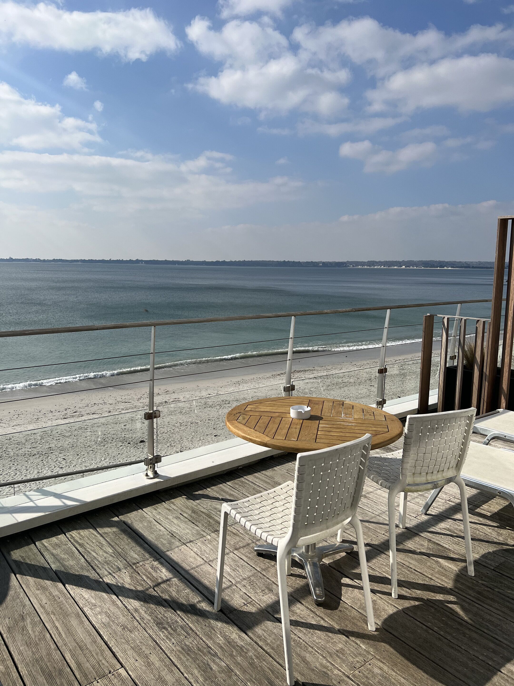 A wooden deck with a round table and white chairs overlooking a beach.