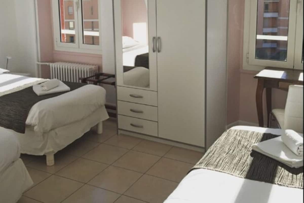 Une chambre avec deux lits, une armoire, un radiateur et une fenêtre donnant sur des immeubles.