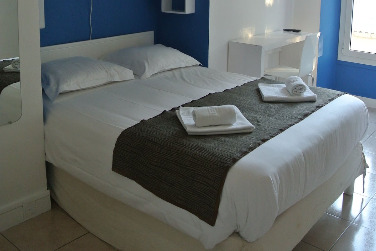 Un lit bien fait, avec des draps blancs, une couverture sombre et une serviette pliée posée sur une table de chevet blanche.