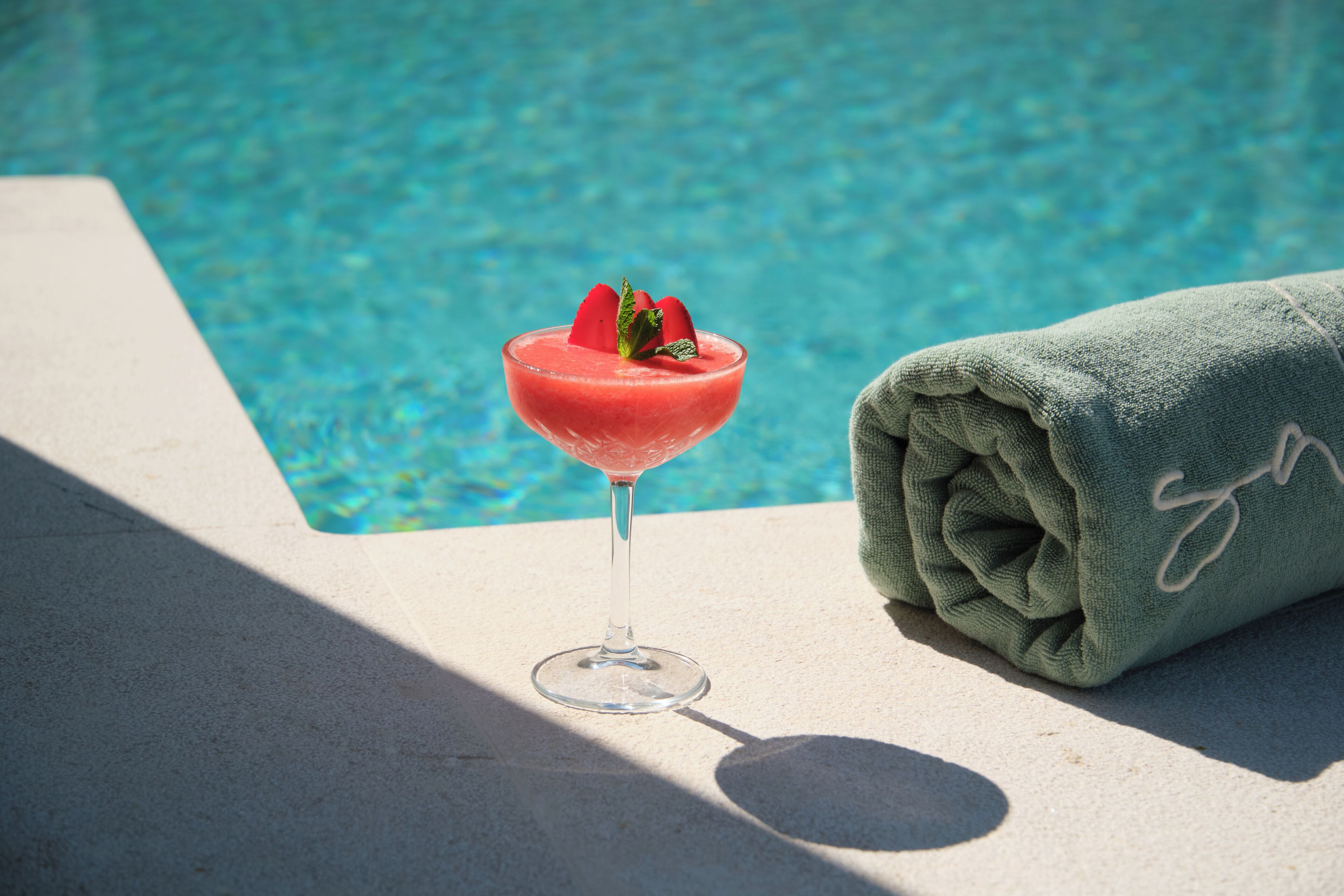 Un cocktail trasparente con una fragola come guarnizione, accanto a un asciugamano arrotolato, vicino a una piscina.