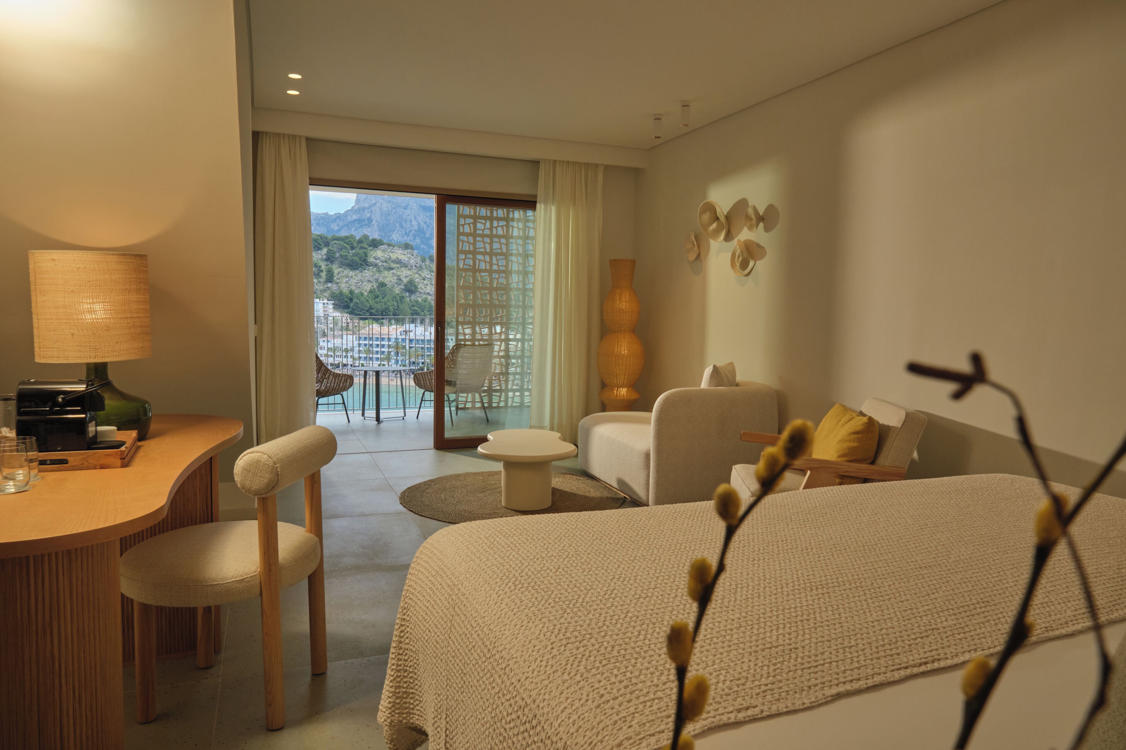 A hotel room with a bed, a desk with a lamp, a chair, a sofa, a small table, and a view of the mountains through a glass door.
