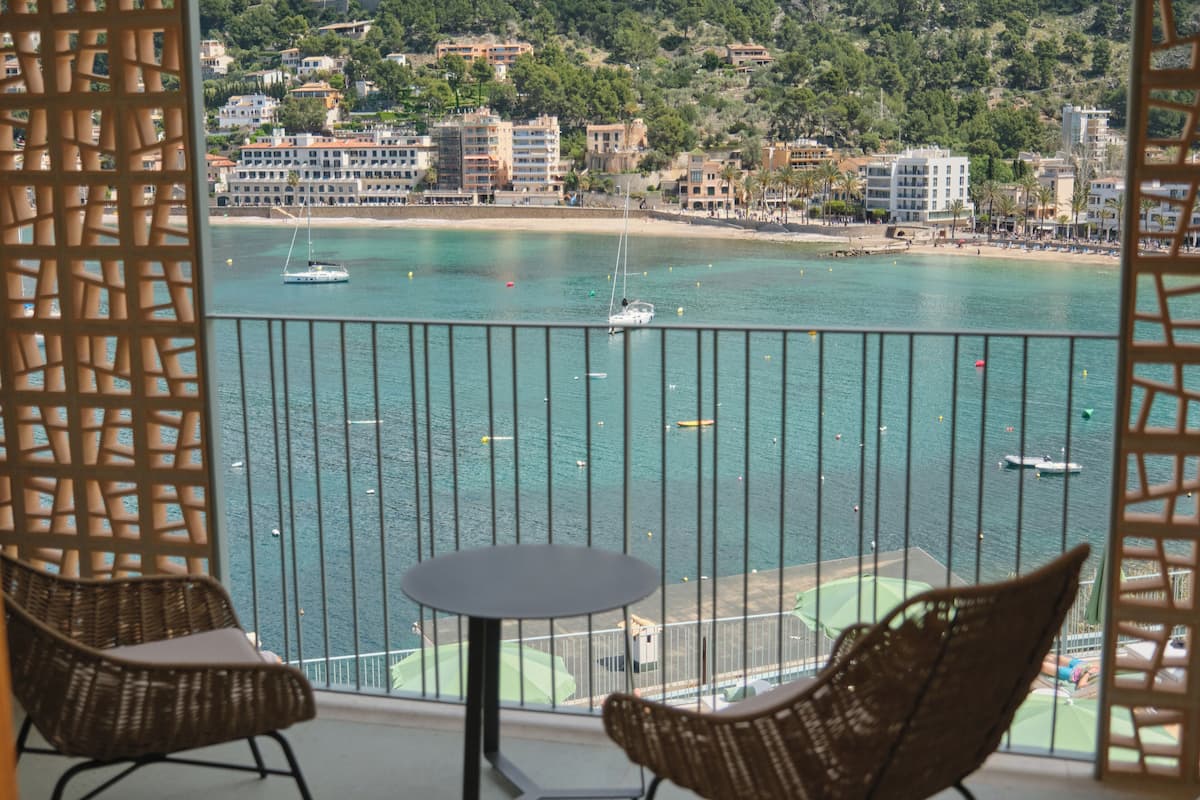 A balcony with wicker chairs and a small table, overlooking a coastal area with buildings and boats.