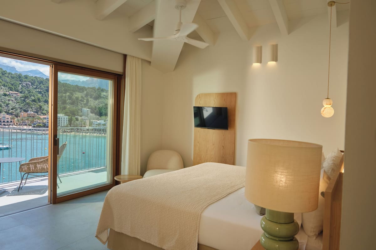 A hotel room with a bed, a chair, a TV, and a balcony with a view of the sea and mountains.