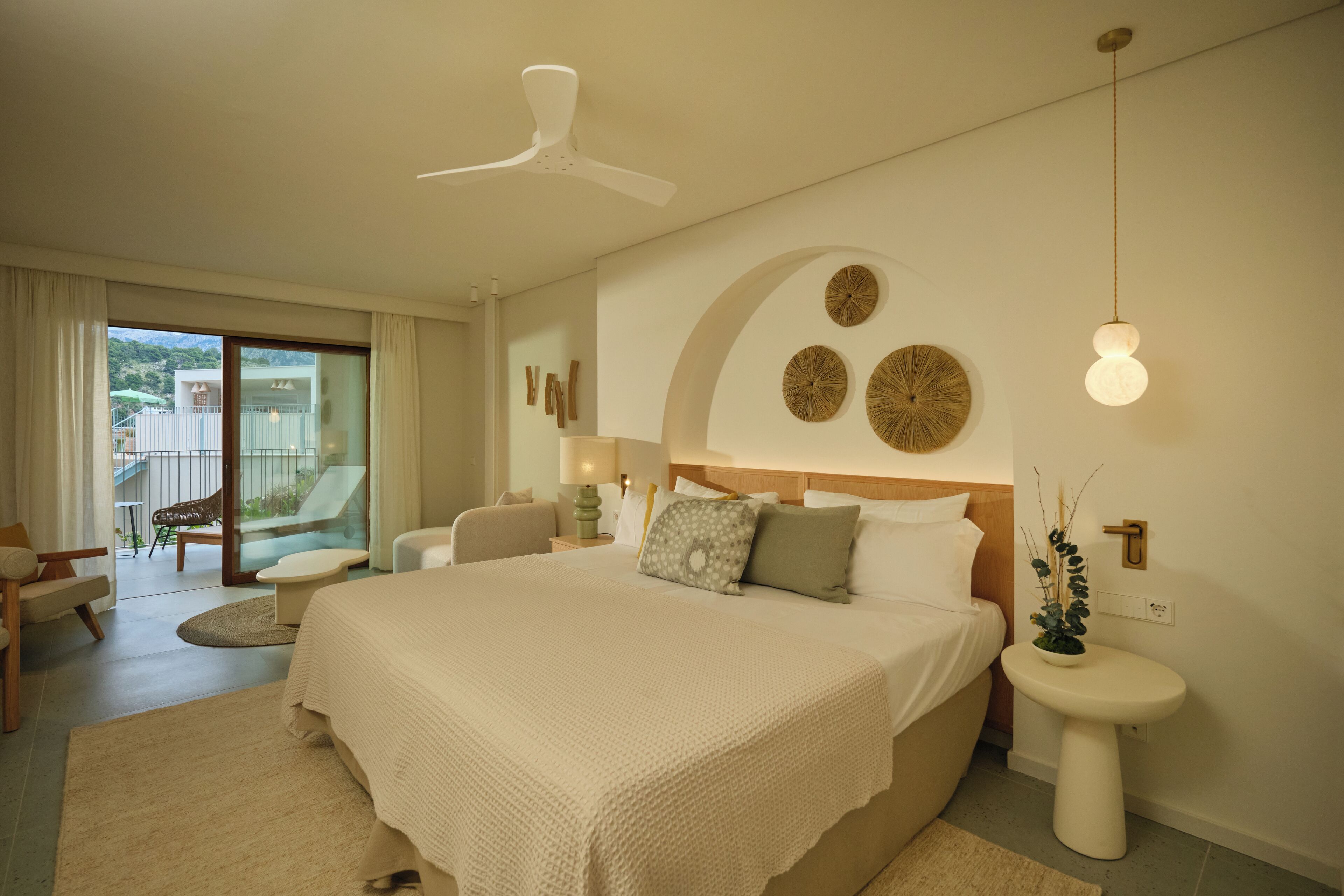 A bedroom with a large bed, a ceiling fan, a bedside table with a lamp, and a view of the outdoors.