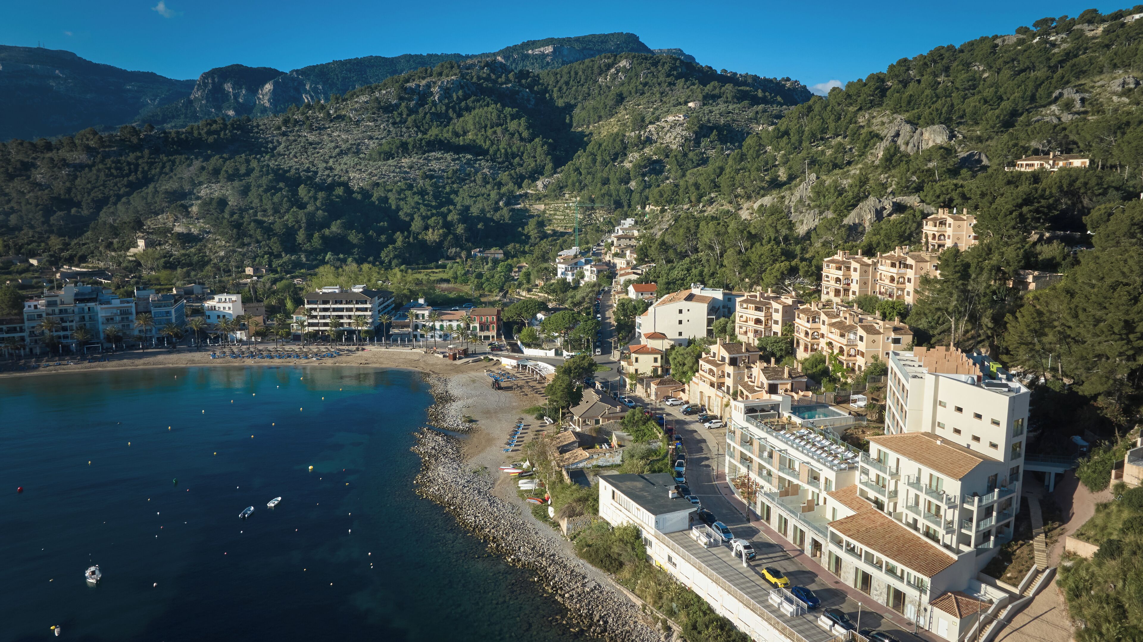 A coastal town with buildings along the beach, a clear blue sea, and mountains in the background.