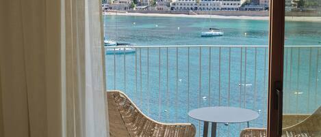A balcony with wicker chairs and a small table, overlooking a coastal town with buildings and boats on the water.