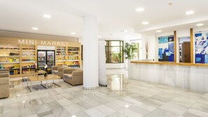 A clean, modern lobby with a reception desk, a seating area with sofas, and a 'Mini Market' with shelves stocked with various products. - Globales Nova (Calvia)
